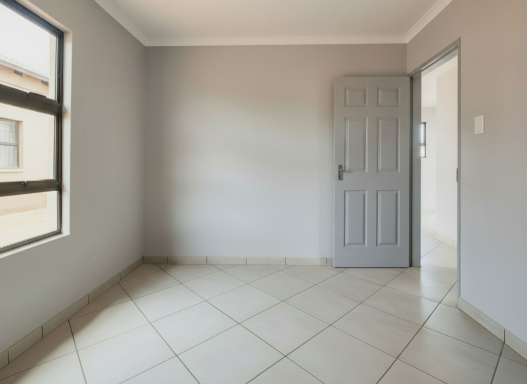 Bedroom in a Windmill Park X32 home showing ceramic tiled floors, an aluminium window, and a grey internal door leading to the main living area.